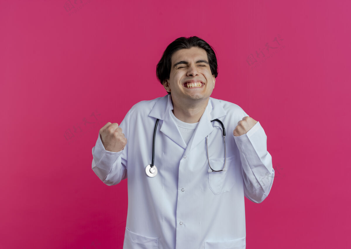 医生快乐的年轻男医生穿着医用长袍和听诊器做“是”的手势 闭上眼睛 隔离在粉红色的墙上 留有复印空间关闭手势年轻