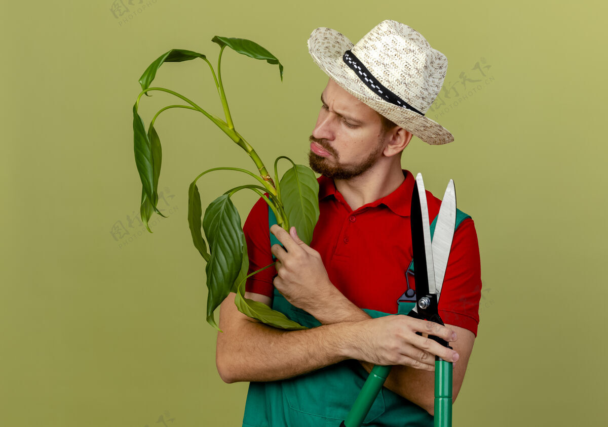 英俊困惑的年轻英俊的斯拉夫园丁穿着制服 拿着帽子 看着橄榄绿墙上孤立的植物花园斯拉夫帽子