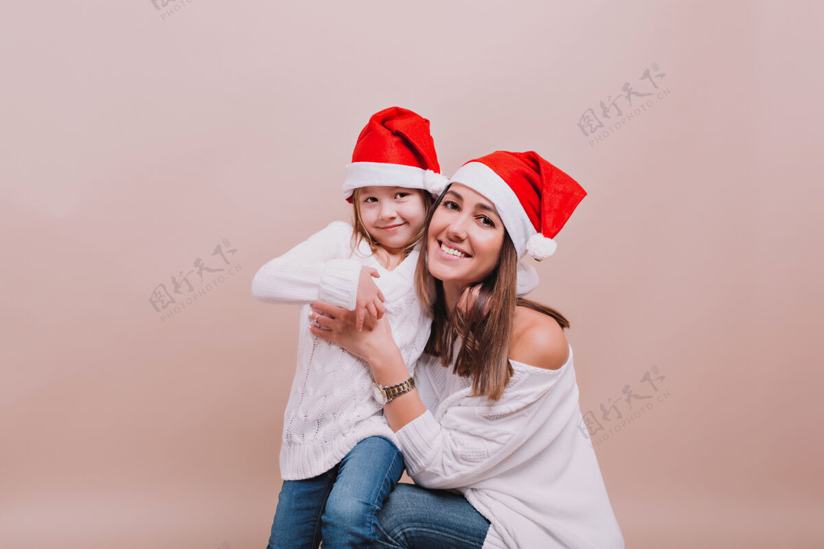 快乐美丽的母亲和小可爱的女儿穿着白色套头衫和圣诞老人的帽子画像节日爱可爱