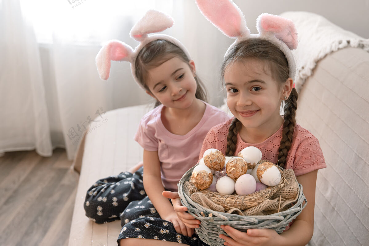 装饰两个滑稽的小女孩在家里的兔子耳朵沙发上与一篮子复活节彩蛋传统兔子房子