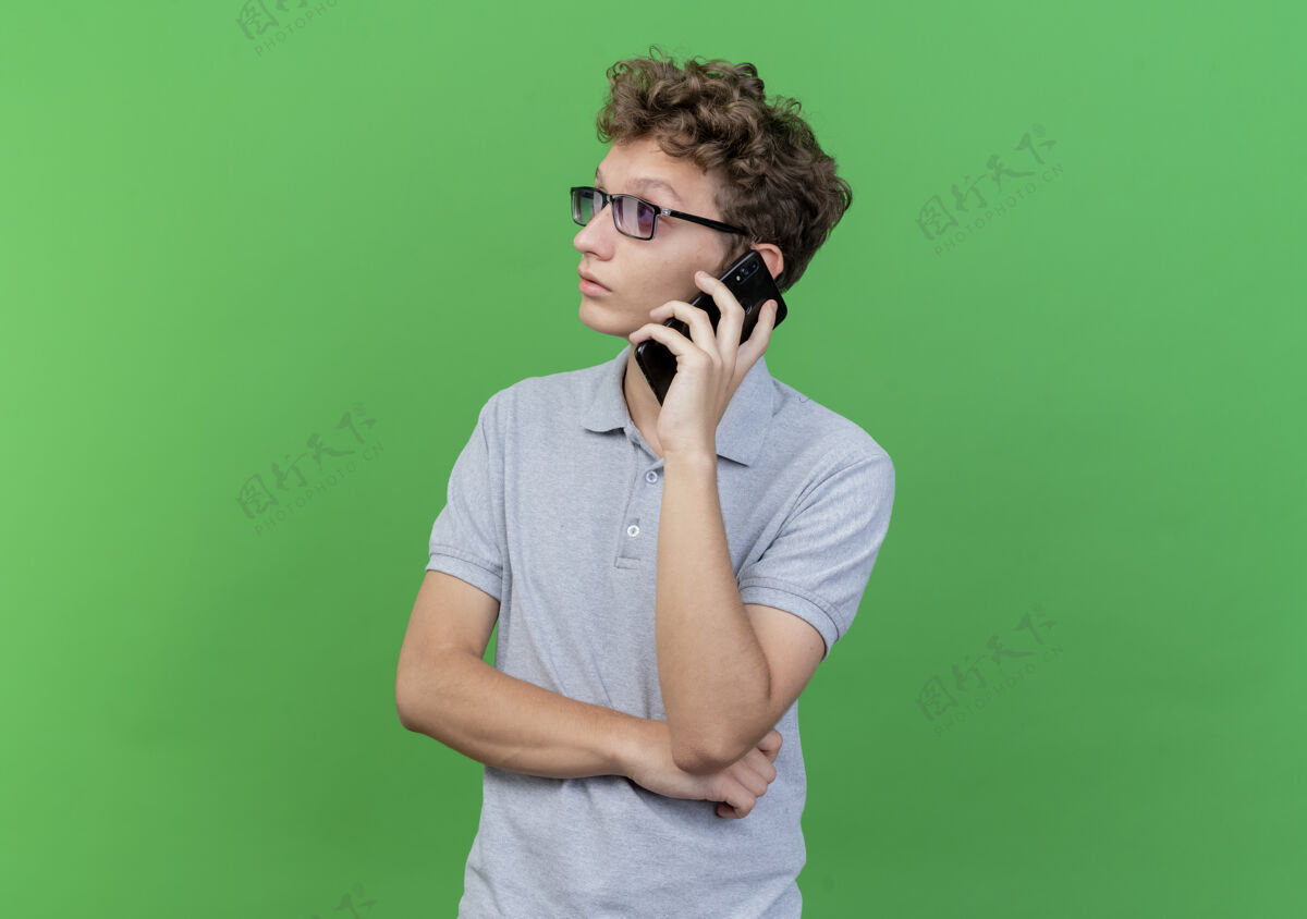 困惑戴着黑眼镜 身穿灰色马球衫的年轻人一边迷茫地看着一旁 一边用绿色手机交谈电话会说话衬衫