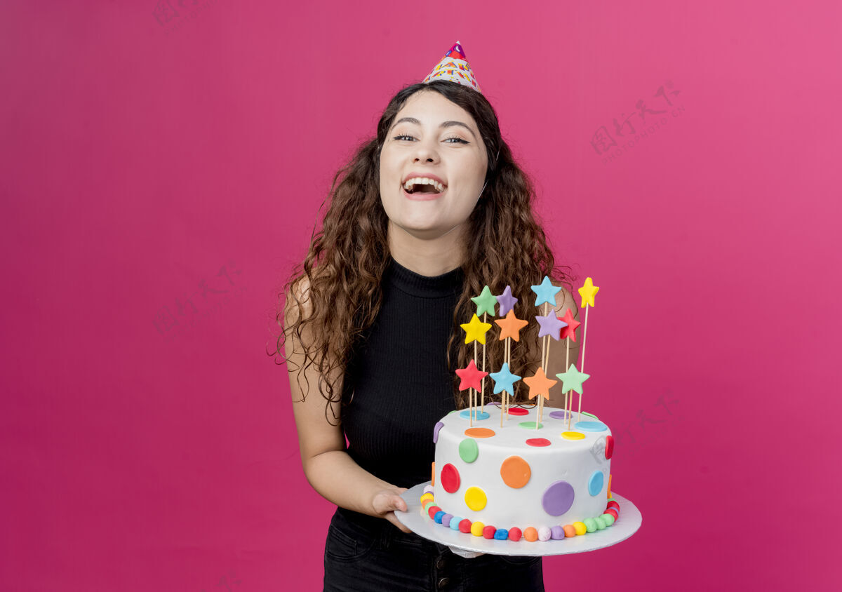 快乐一位年轻漂亮的女士 卷发 戴着节日帽 手里拿着生日蛋糕 开心而兴奋地笑着 生日派对的概念超过了粉色欢呼抱着生日