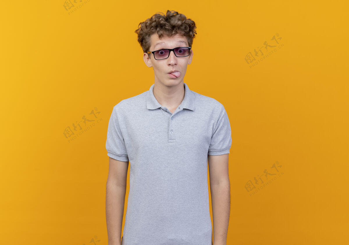 穿戴着黑眼镜的年轻人穿着灰色马球衫快乐而积极地伸出舌头遮住橙色站年轻灰色
