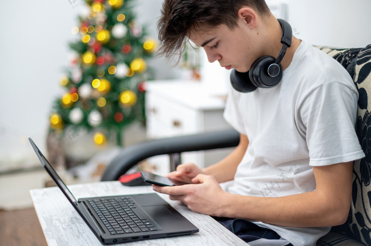 便携十几岁的男孩正在家里用带耳机的智能手机 膝盖上放着笔记本电脑墙上挂着圣诞树严肃而专注的脸儿童媒体肖像