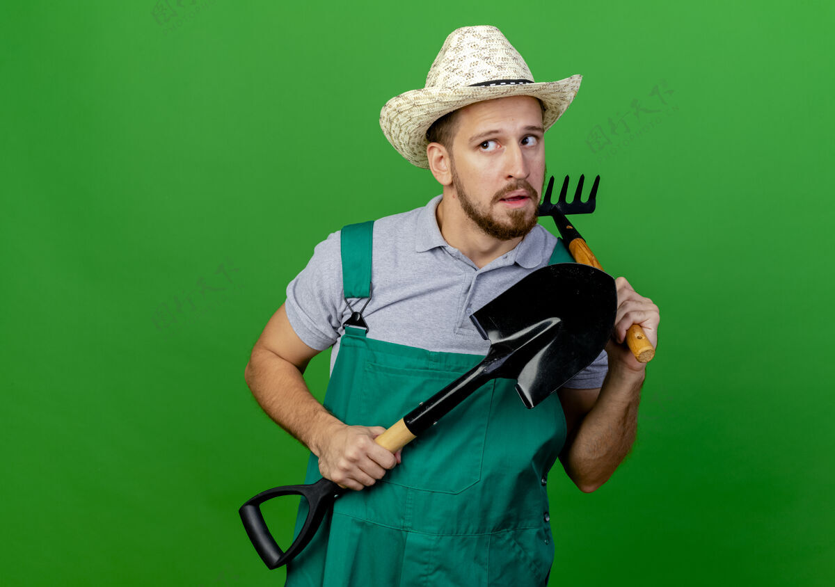 黑桃年轻英俊的斯拉夫园丁 穿着制服 戴着帽子 手里拿着铲子和耙子 看着旁边绿色帽子花园