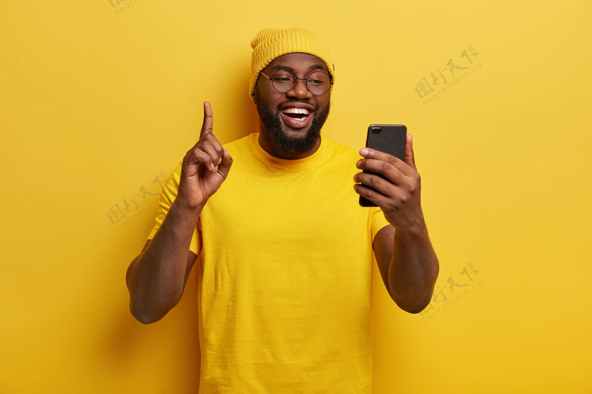 眼镜快乐丰满的男人举起食指 拿着手机 享受闲暇时间上网 戴着黄色帽子和休闲t恤眼镜科技小伙子