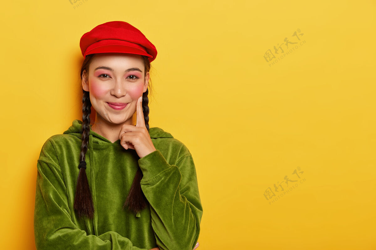 表情快乐的亚洲女孩把食指放在脸颊上 开心地看着镜头 脸颊上涂着胭脂 头戴红色贝雷帽 穿着绿色天鹅绒运动衫女人时尚美丽