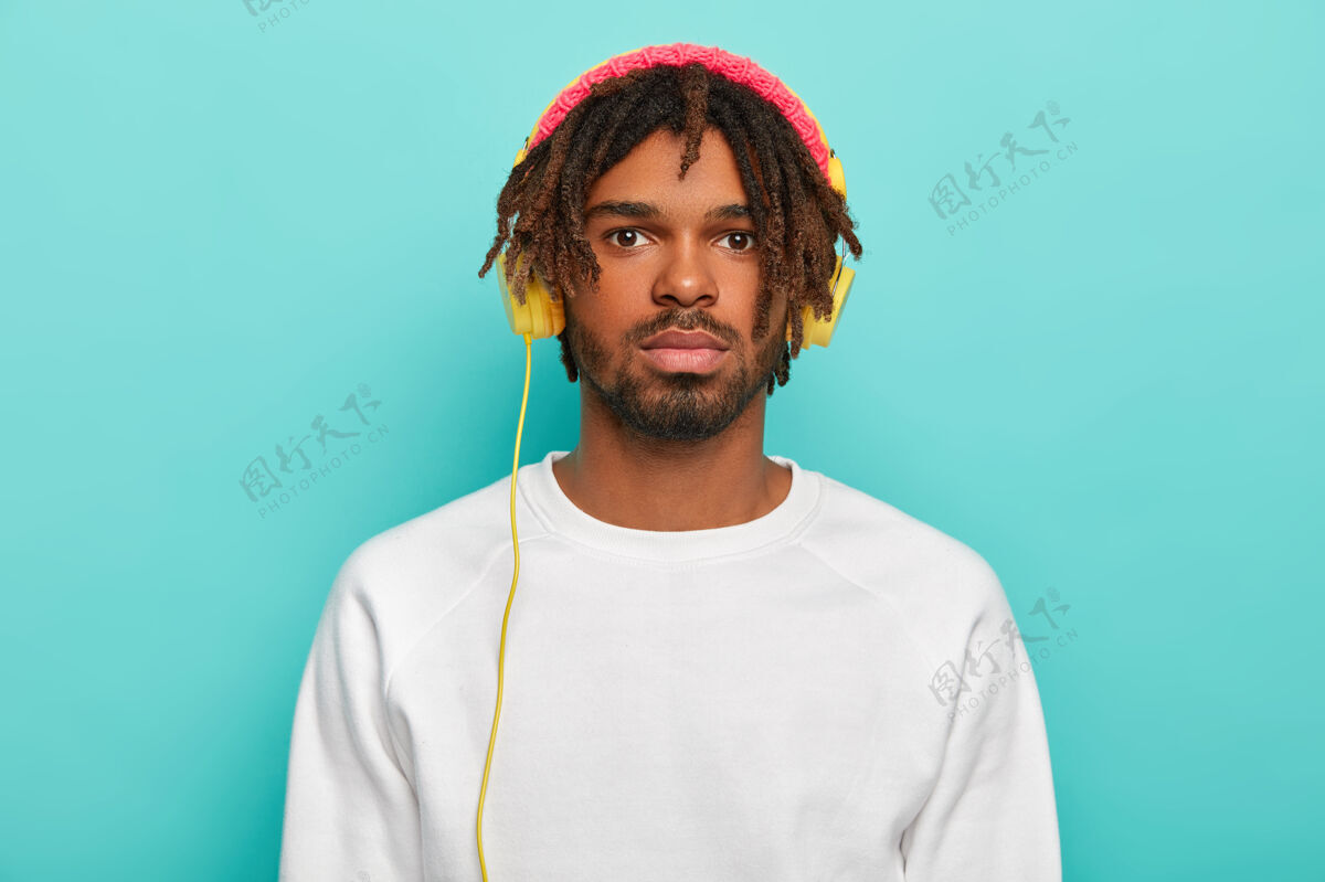 帽子帅气自信的男性 戴着长发锁 戴着耳机 听着温暖的音乐或收音机 享受着舒适的氛围 穿着白色休闲毛衣 戴着玫瑰色的头饰科技站听