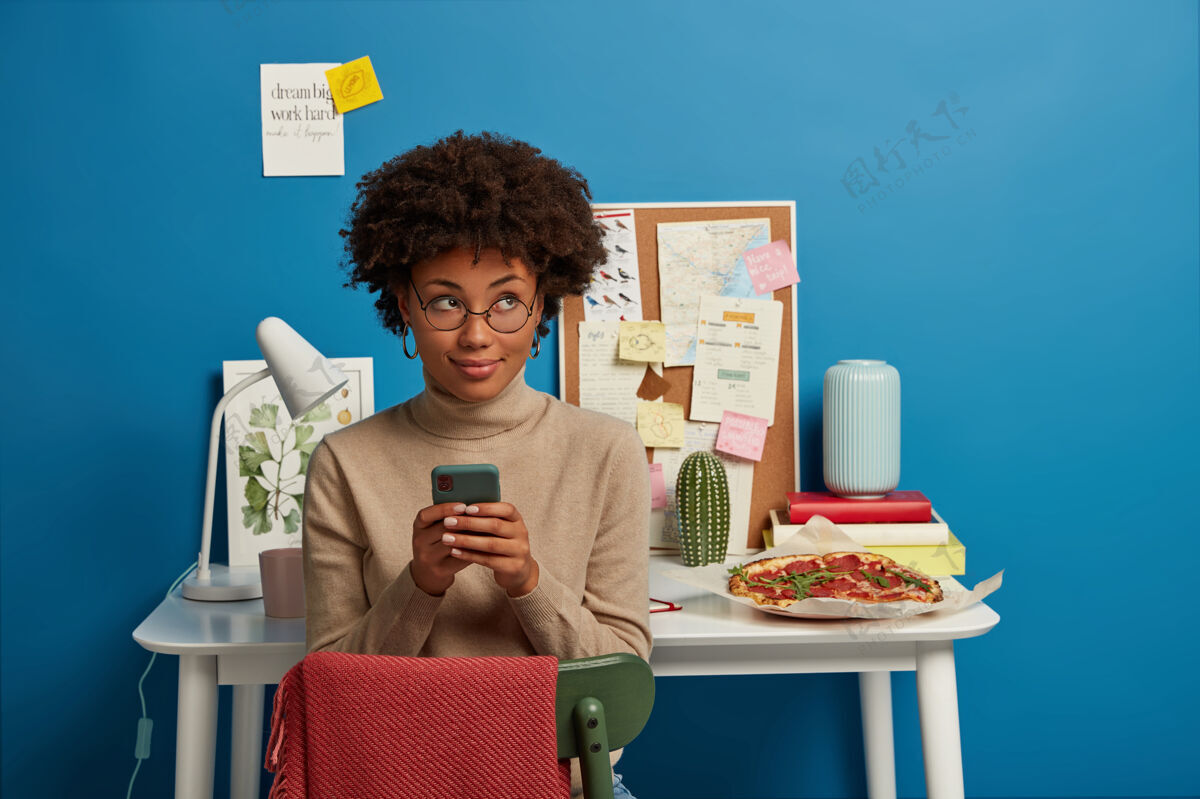 休闲迷人的卷发女性在家远程工作 手里拿着手机 发短信 戴着眼镜 在自己舒适的办公室里摆姿势通信手机桌子
