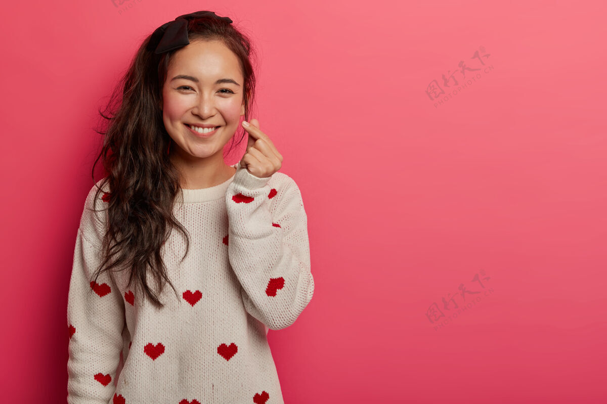 跳投浪漫的中国女人双手合十展示韩国人的心形标志 开心地微笑着承认爱情 表达爱意 穿着印有心形图案的毛衣 隔离在粉色的工作室墙上人人类积极