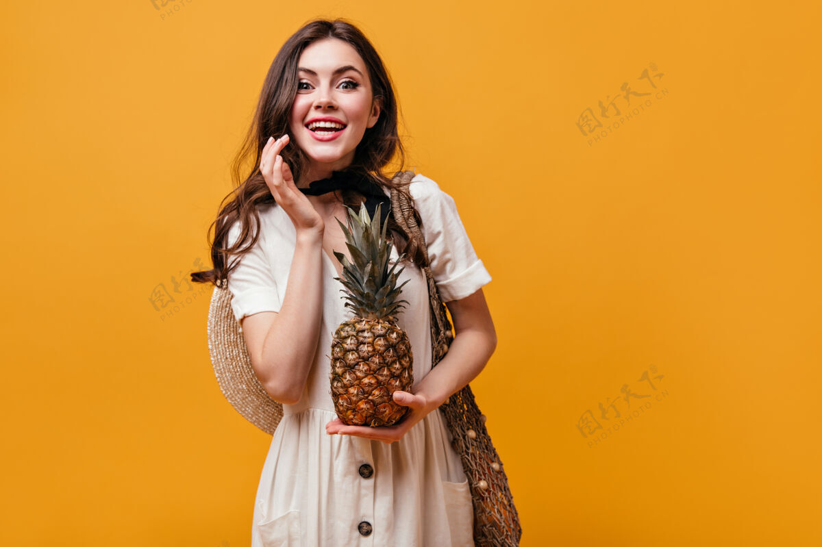 水果绿眼睛的白裙女孩手里拿着菠萝女人笑着拿着橙色背景的购物袋摆姿势包女性化妆品