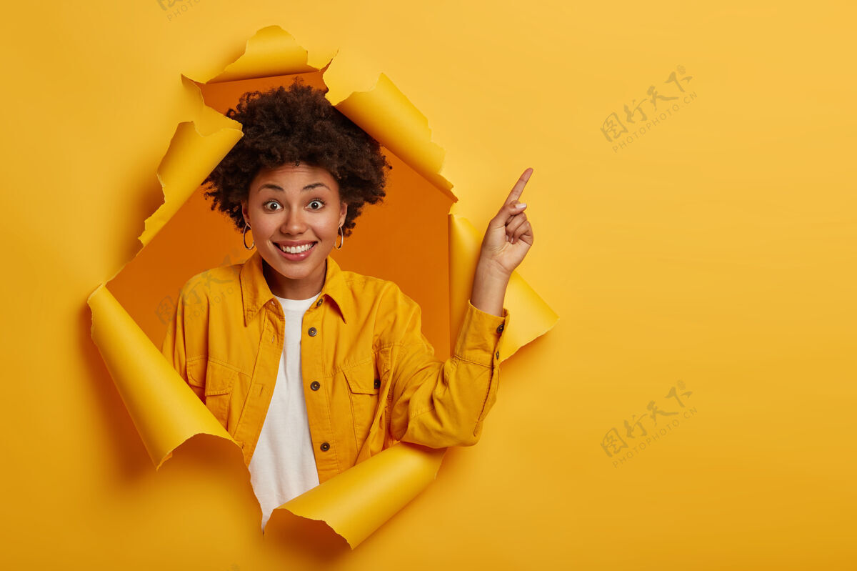 年轻正面非裔美国女性的摄影棚镜头指着上面的复制空间 被好消息所激动 笑容可掬 穿着黄色夹克 站在撕破的纸洞里方向内容女性