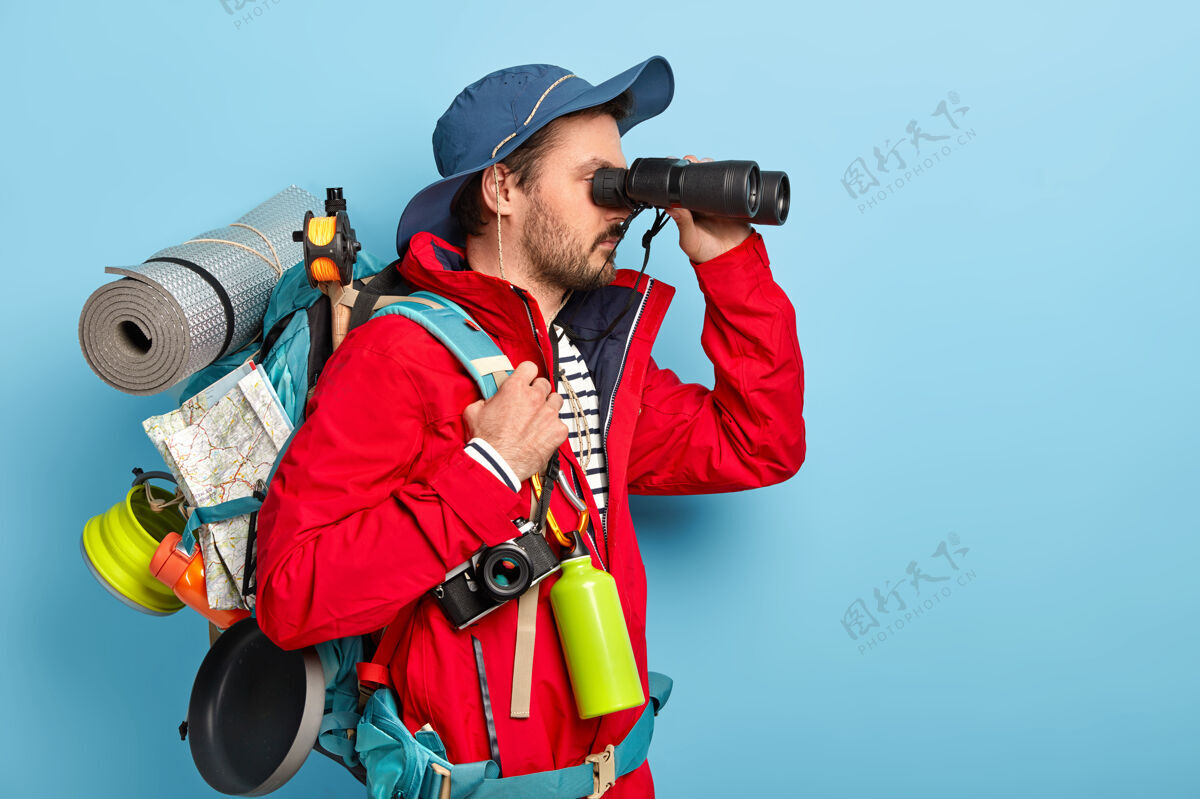 休闲男探险家穿着休闲装 把望远镜放在眼睛附近 戴着帽子和夹克 在山里远足徒步旅行背包胡茬
