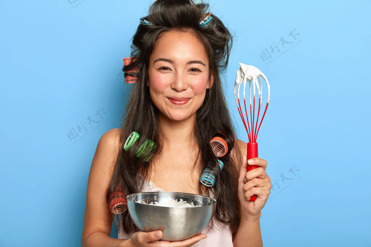 厨师满意的黑发女人在碗里搅拌酸奶油 戴着卷发器混合高兴睡衣