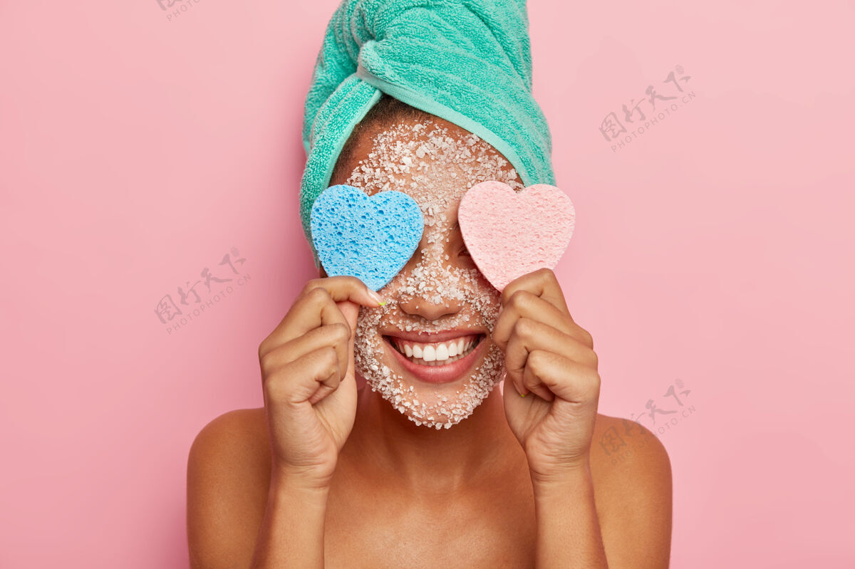 高兴积极的女人在美容过程中很有乐趣 眼睛上贴着两块心形海绵 笑容灿烂 露出洁白的牙齿 头上裹着毛巾 裸体在室内摆姿势人过程水疗
