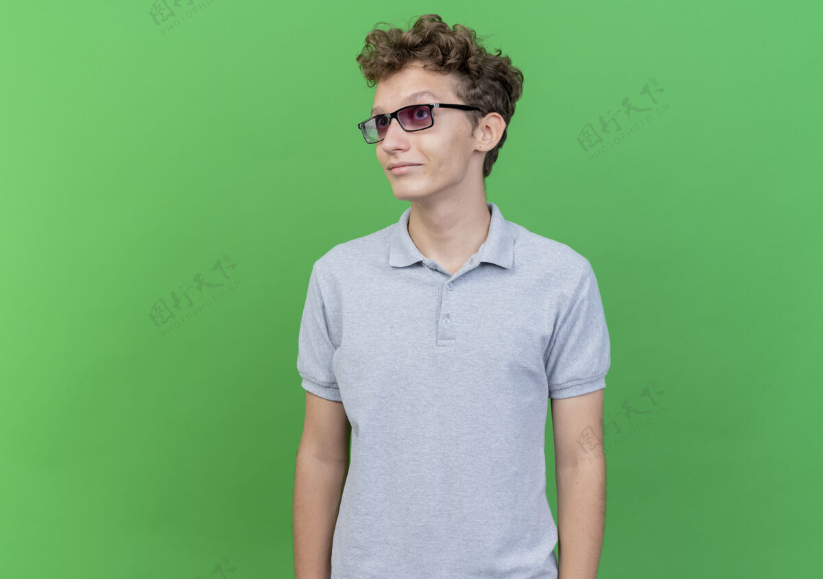 放一边一个戴着黑眼镜 穿着灰色马球衫的年轻人站在绿色的墙上 脸上带着微笑 望向一边微笑眼镜马球