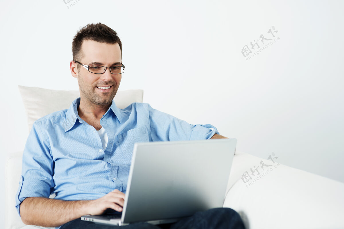 肖像戴眼镜的帅哥在沙发上用笔记本电脑表达积极牙齿微笑上网