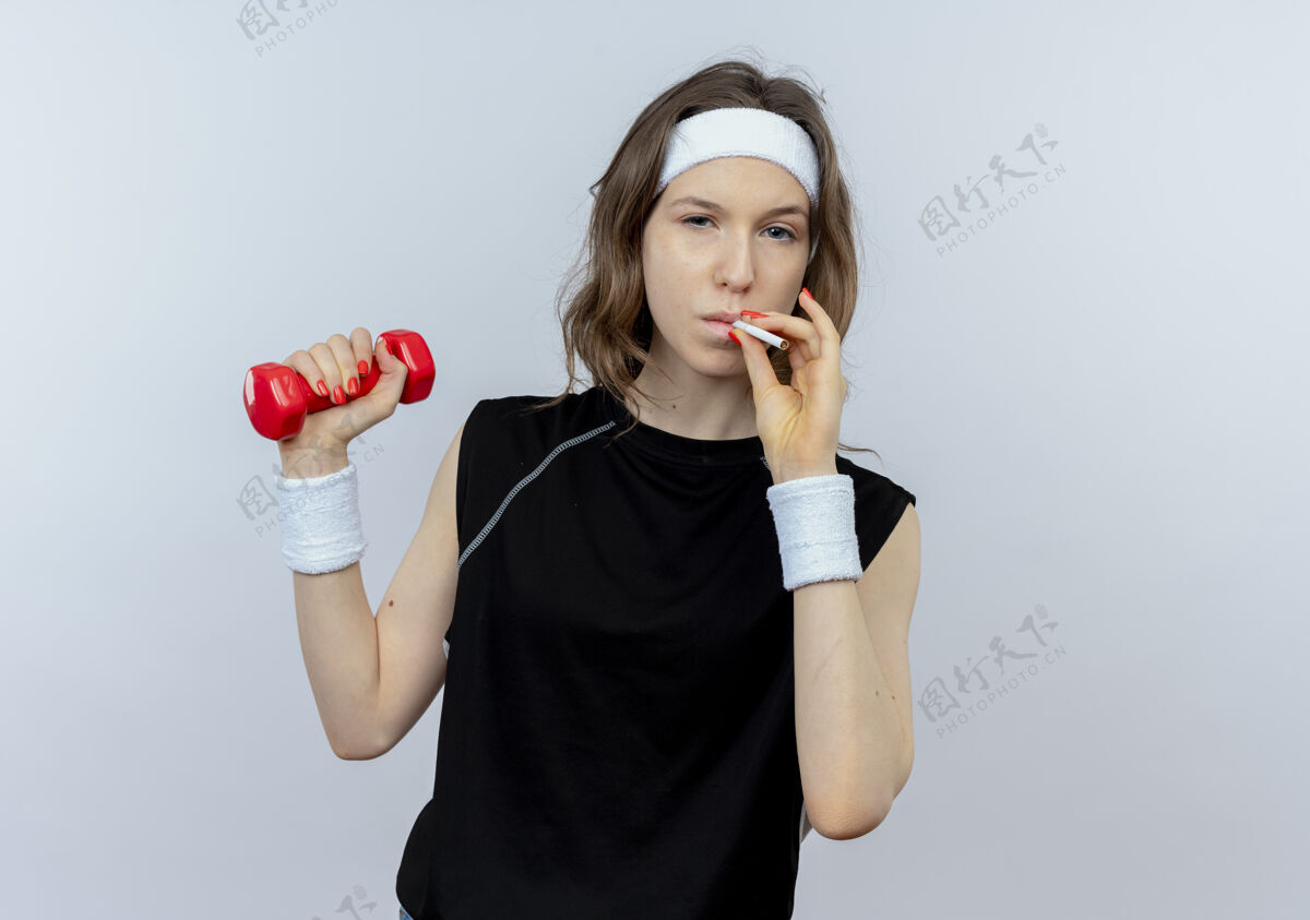 女孩身穿黑色运动服 头戴哑铃 吸烟的年轻健身女孩站在白色墙壁上锻炼身体习惯工作年轻