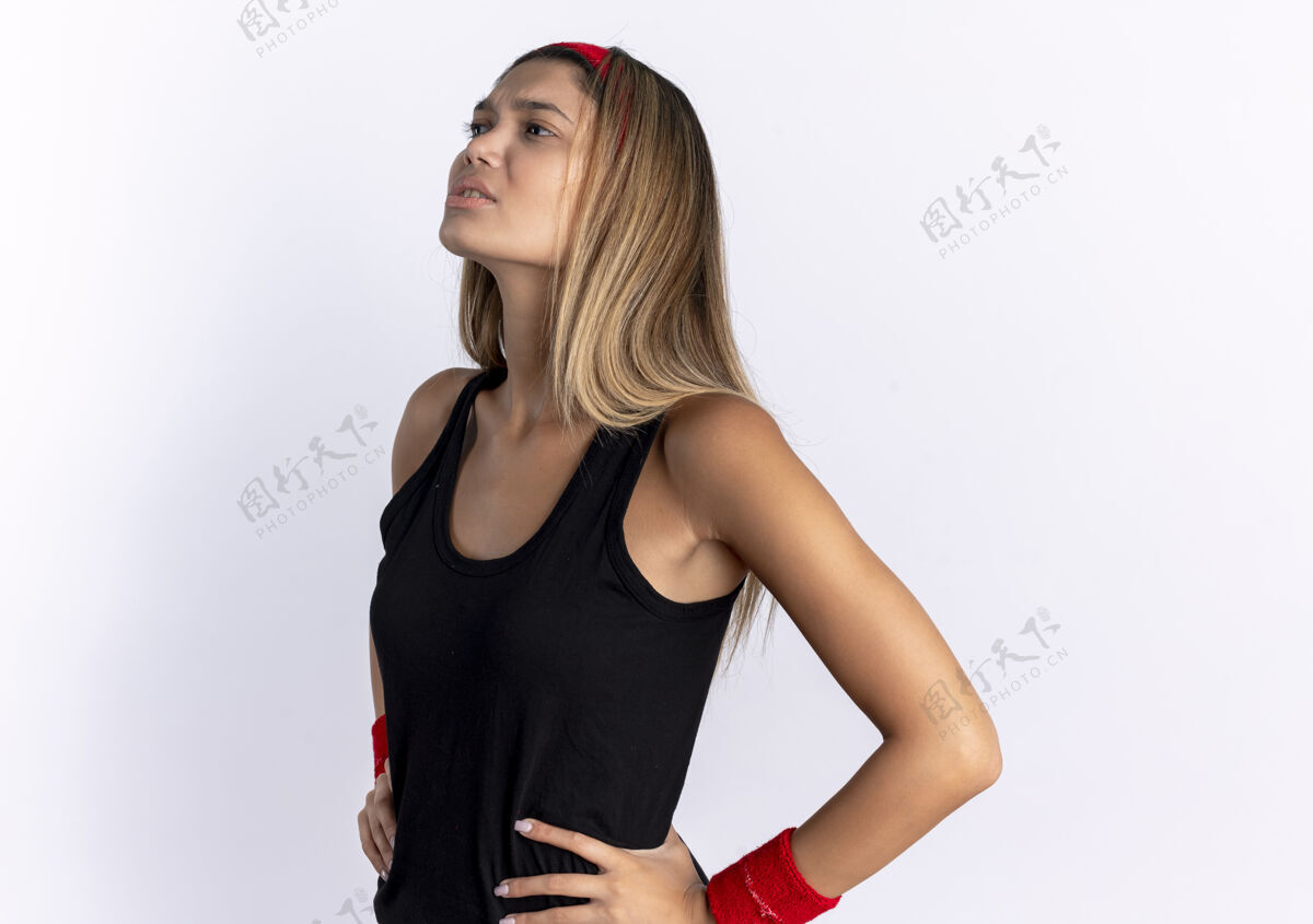 女孩身穿黑色运动服 头戴红色头巾的年轻健身女孩站在白色的墙上 看上去很困惑年轻头带站