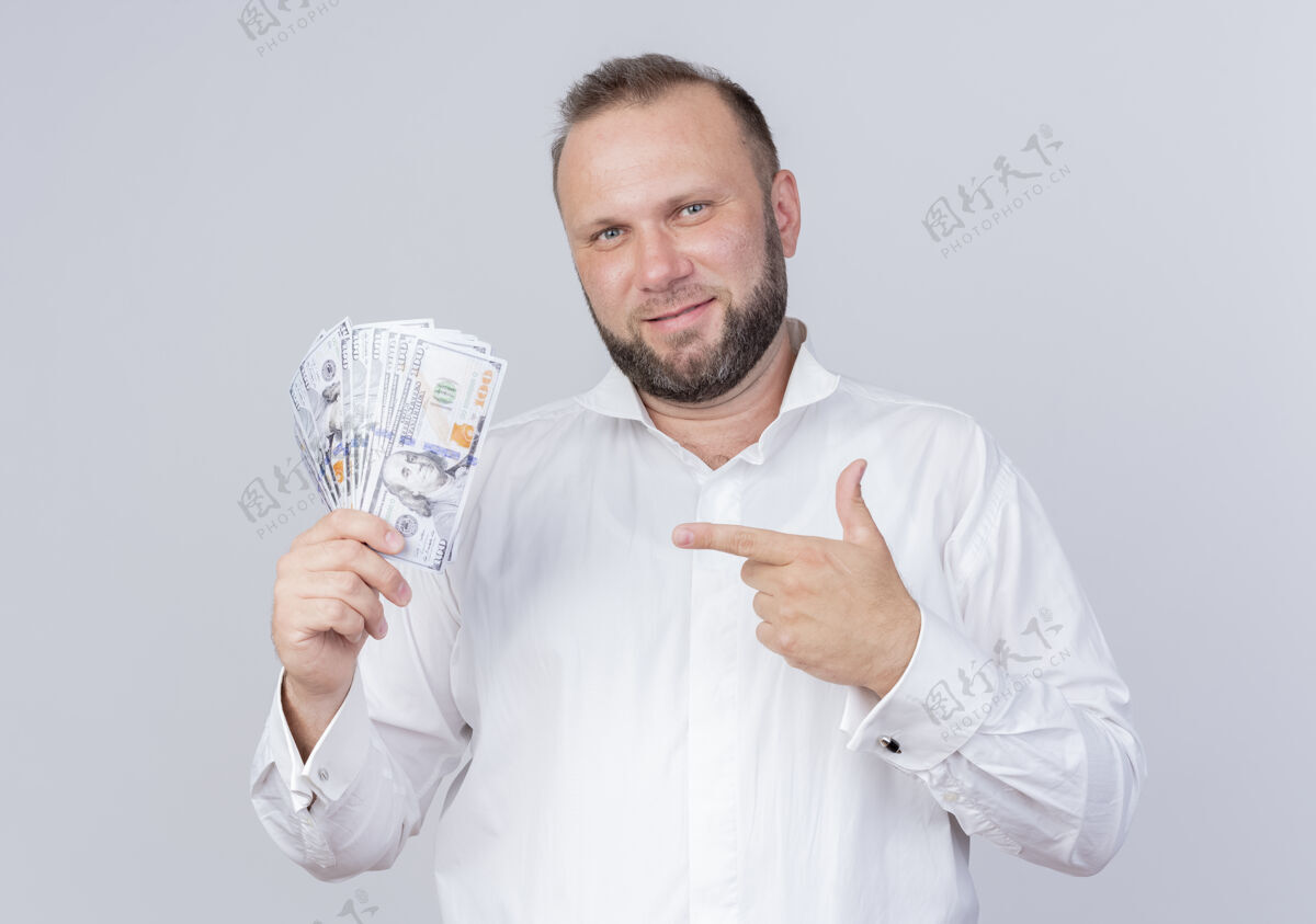 胡子一个留着胡子的男人穿着白衬衫 手里拿着现金 用食指指着钞票 自信地站在白墙上微笑着男人钱戴着