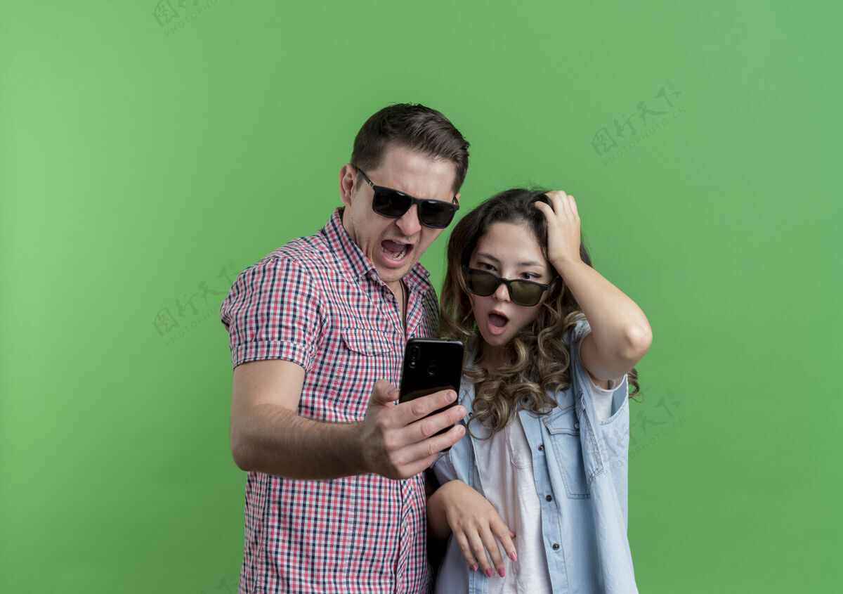 立场一对穿着休闲服 戴着黑眼镜的年轻夫妇站在绿色的墙上 看着自己的智能手机屏幕 既惊讶又困惑女人衣服穿着