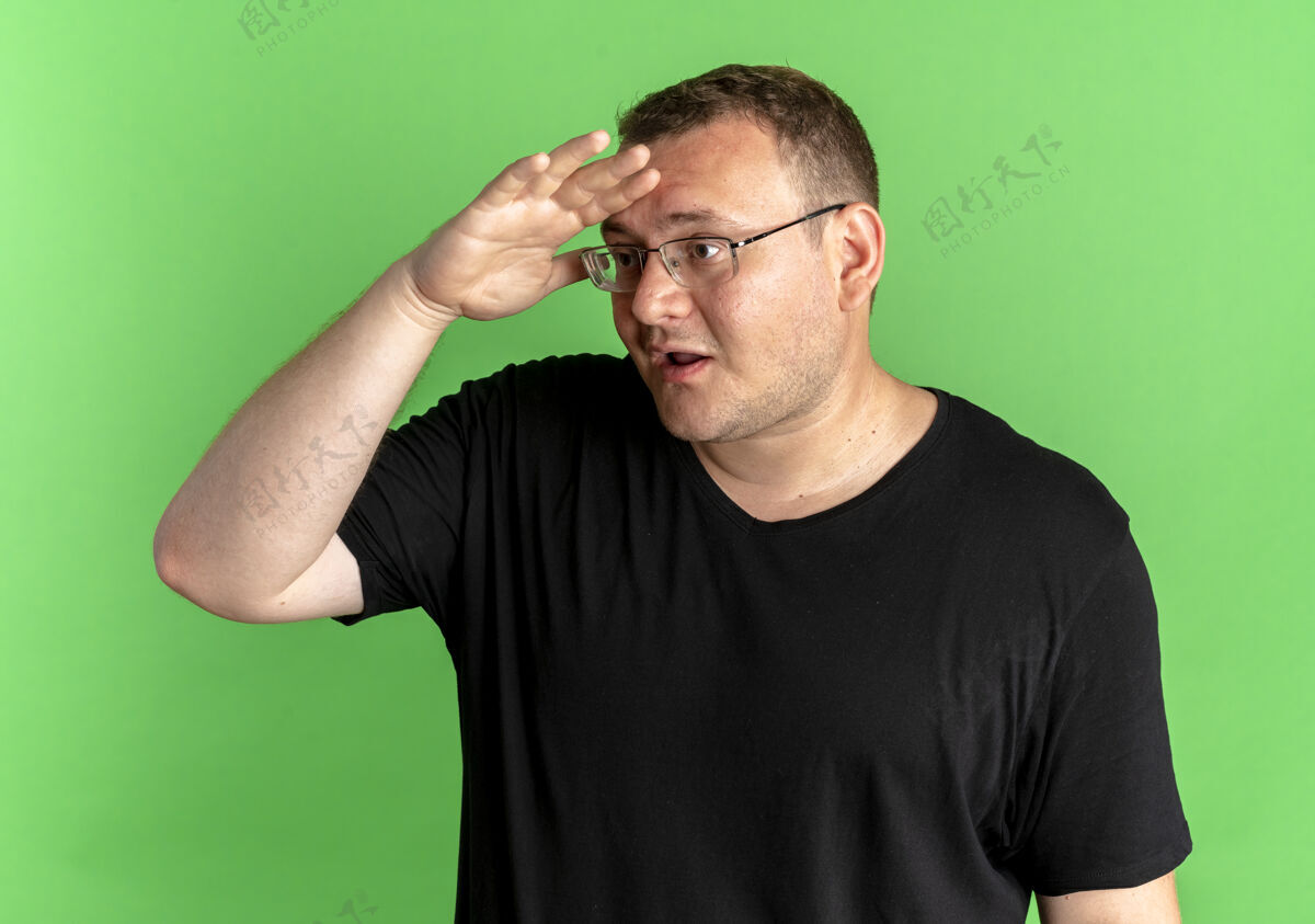 超重一个戴眼镜的胖男人穿着黑色t恤望着远处 手捂着额头惊讶地站在绿色的墙上眼镜额头远离