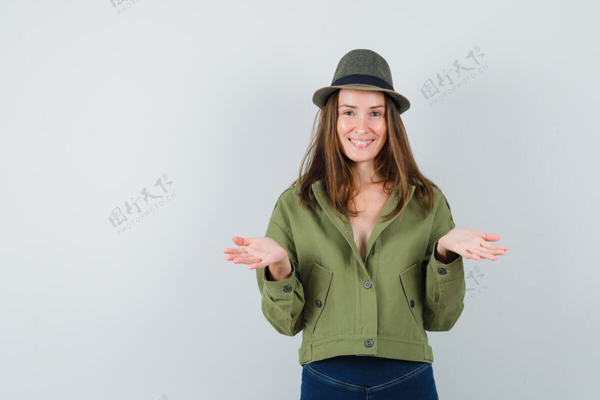 黑发年轻女性假装拿着或展示夹克 裤子 帽子里的东西 看起来很高兴正面图假装肖像微笑