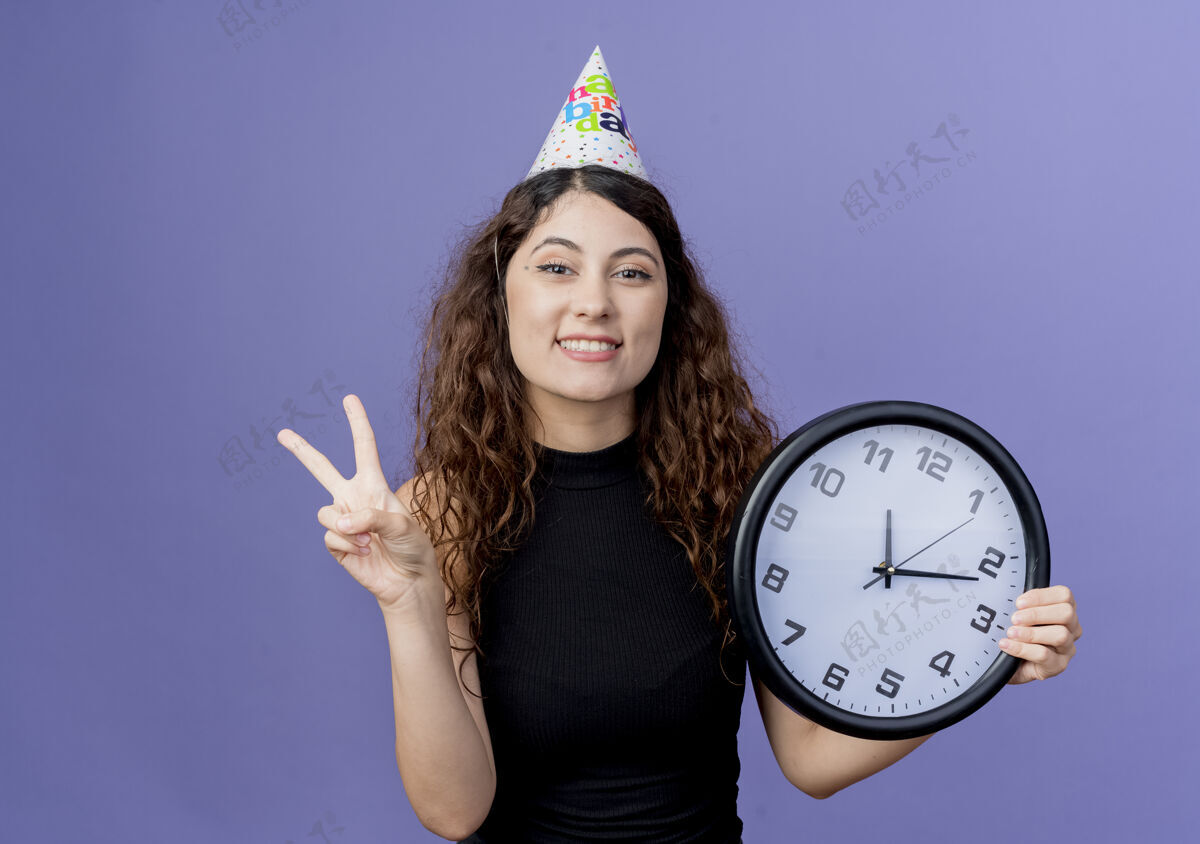 抱着一位年轻漂亮的女士 卷发 头戴节日帽 手持挂钟 微笑着 站在蓝色的墙上 展示v字生日派对的概念微笑表演年轻