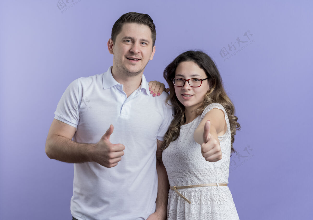 站一对年轻夫妇站在蓝色的墙上 微笑着竖起大拇指看着摄像机微笑情侣秀
