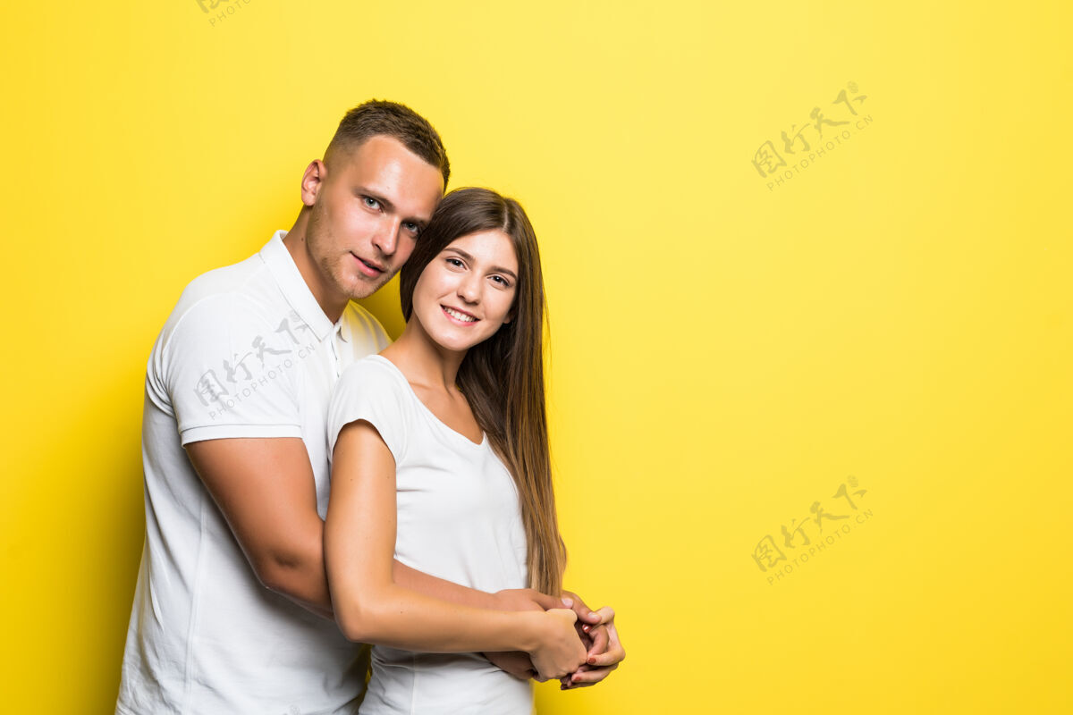 脸一对身着白色t恤的幸福年轻夫妇在黄色背景上拥抱乐趣拥抱关系