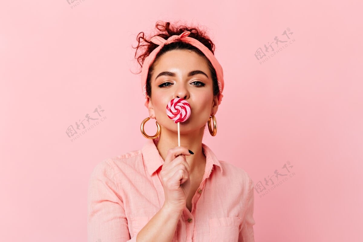 发型蓝眼睛卷曲的女孩 戴着粉色的头带和巨大的金耳环 在粉色的空间里吃糖果红色唇膏糖果正品