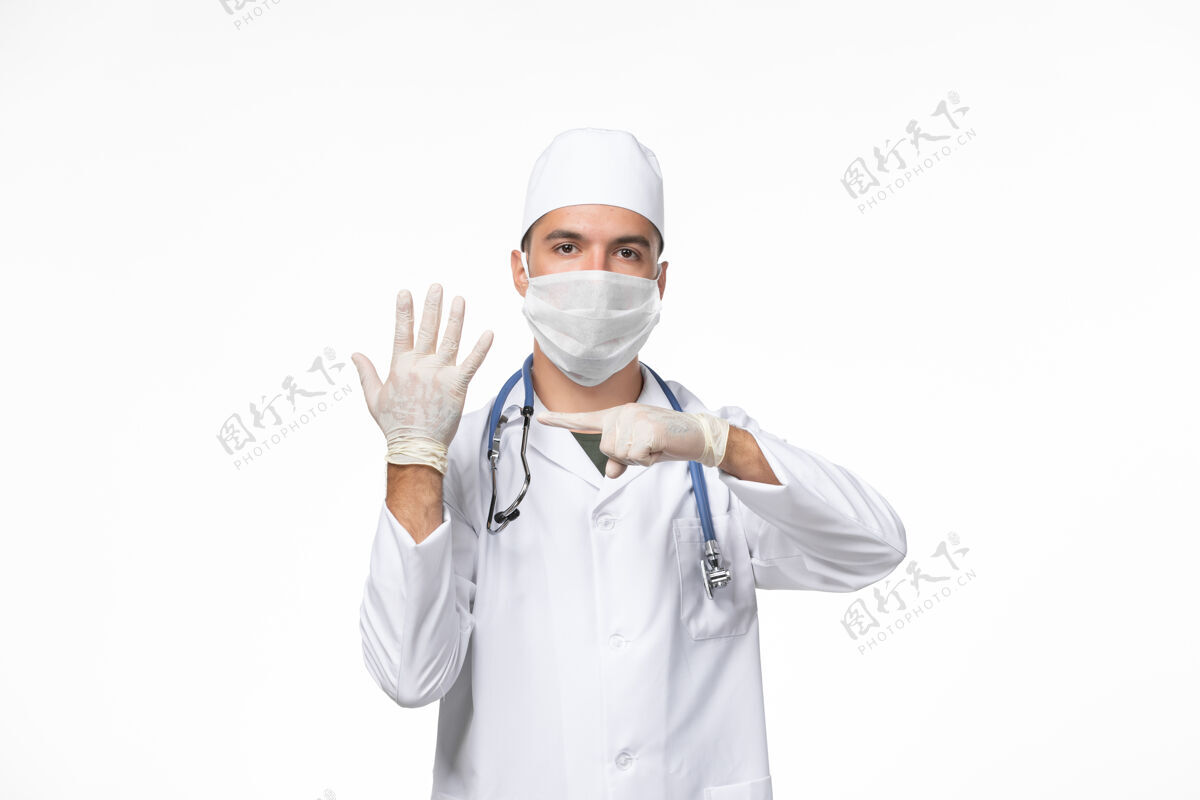 病毒正面图：男医生穿着医疗服 戴着口罩 用听诊器对着白色办公桌上的柯维病大流行病毒制服专业听诊器