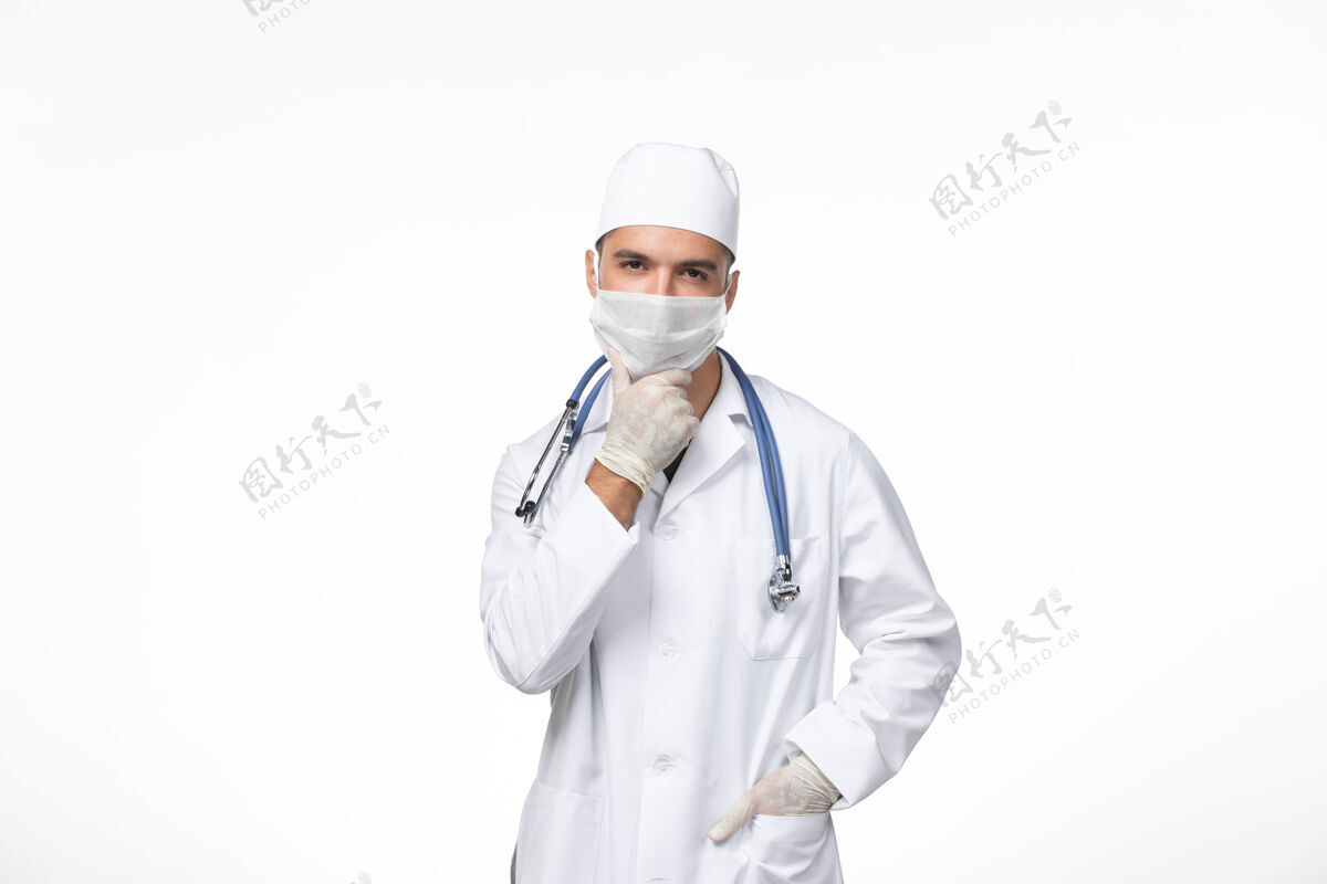医疗正面图男医生穿着医疗服 戴着防冠状病毒口罩 用蓝色听诊器思考白墙冠状病毒-疾病大流行病毒专业视图正面