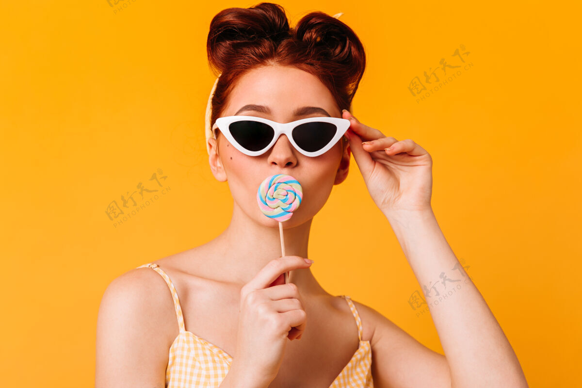 时尚戴着太阳镜舔着硬糖的好女人黄色空间里孤立着一个戴着棒棒糖的小女孩的前视图糖果甜蜜发型