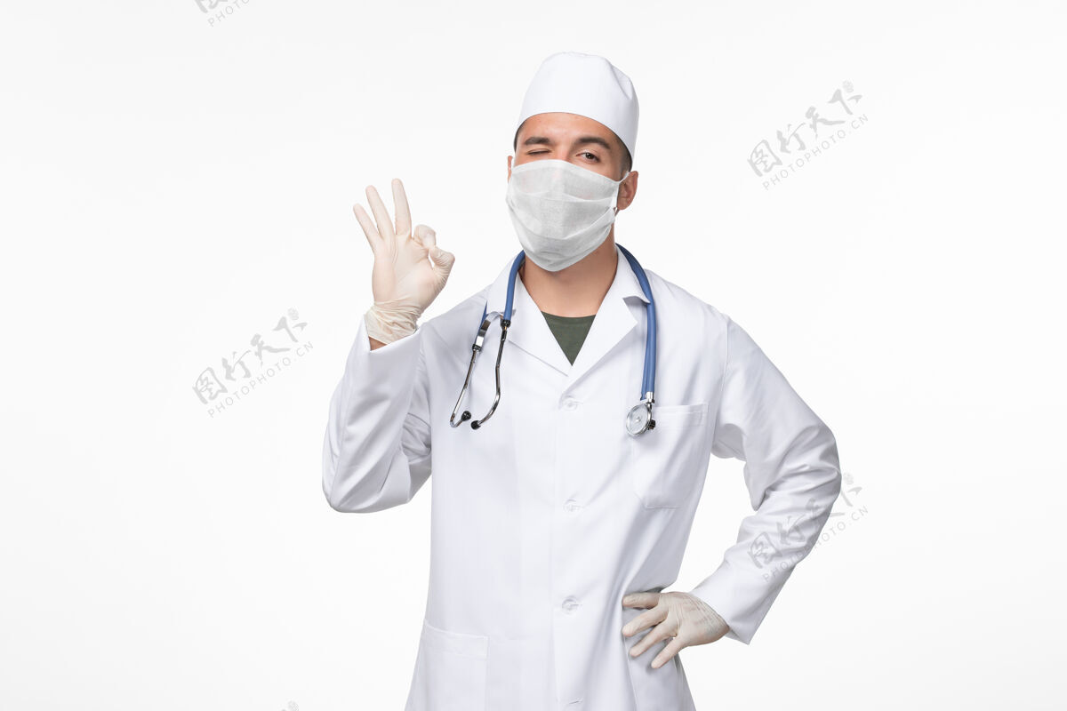 医疗正面图：男医生穿着医疗服 戴着口罩 戴着听诊器 对着白墙病毒-病毒-疾病大流行眨眼面具病毒佩戴