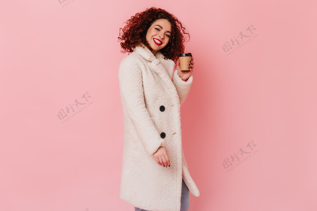 漂亮可爱的黑发女人 穿着浅羊毛外套 带着雪白的微笑 在粉色的空间里捧着一杯硬纸板咖啡卷曲红色嘴唇女性