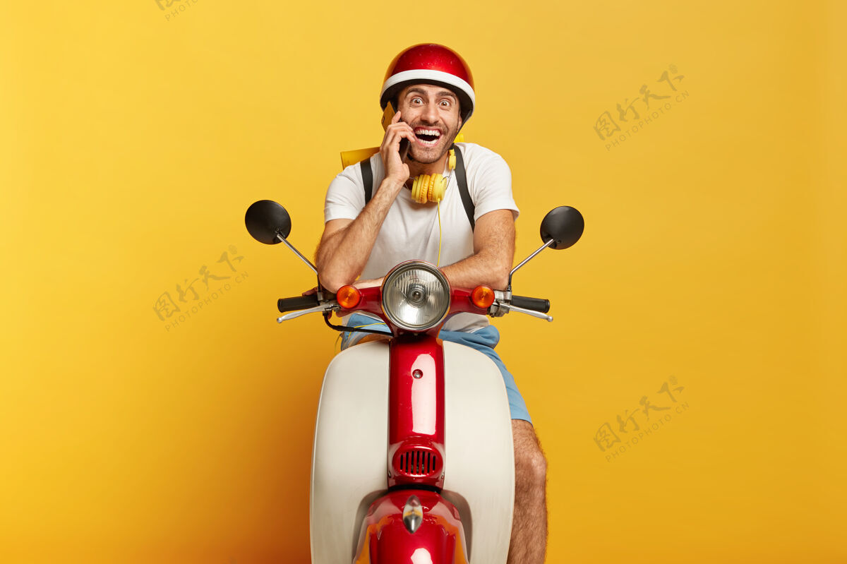 牙齿快乐的摩托车手在自己的快速交通工具上摆姿势 通过智能手机给客户打电话 长途旅行 戴头盔 脖子上戴着立体声耳机 对着镜头微笑男司机开着摩托车摩托车摩托车手欣喜若狂