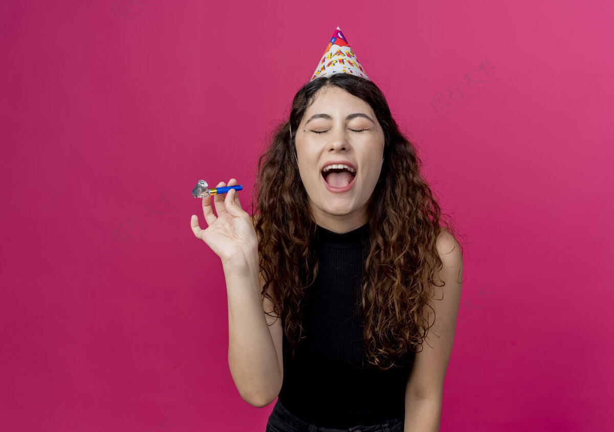 卷发年轻漂亮的女人卷发戴着节日帽疯狂的生日快乐派对概念站在粉红色的墙上疯狂站起来年轻