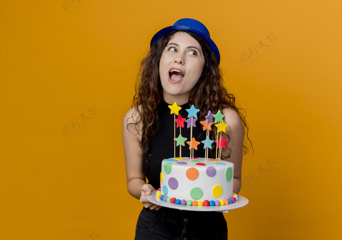 女人一个年轻漂亮的女人 卷发 戴着节日帽 手里拿着生日蛋糕 站在橘色的墙上 又高兴又惊讶蛋糕帽子抱着
