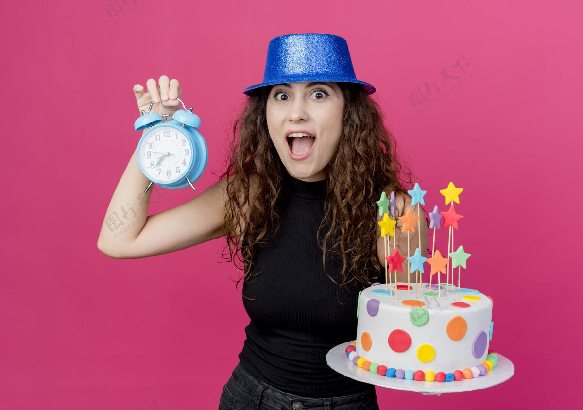 站着一位年轻漂亮的女士 卷发 戴着节日礼帽 手里拿着生日蛋糕和闹钟 站在粉色的墙上 看起来很惊讶生日派对的概念抱着女人蛋糕