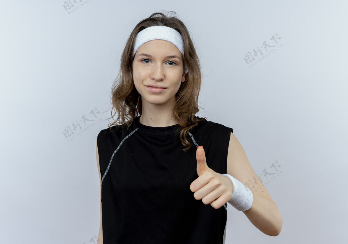 年轻身穿黑色运动服的年轻健身女孩站在白色的墙上 竖起大拇指微笑着健康运动装女孩
