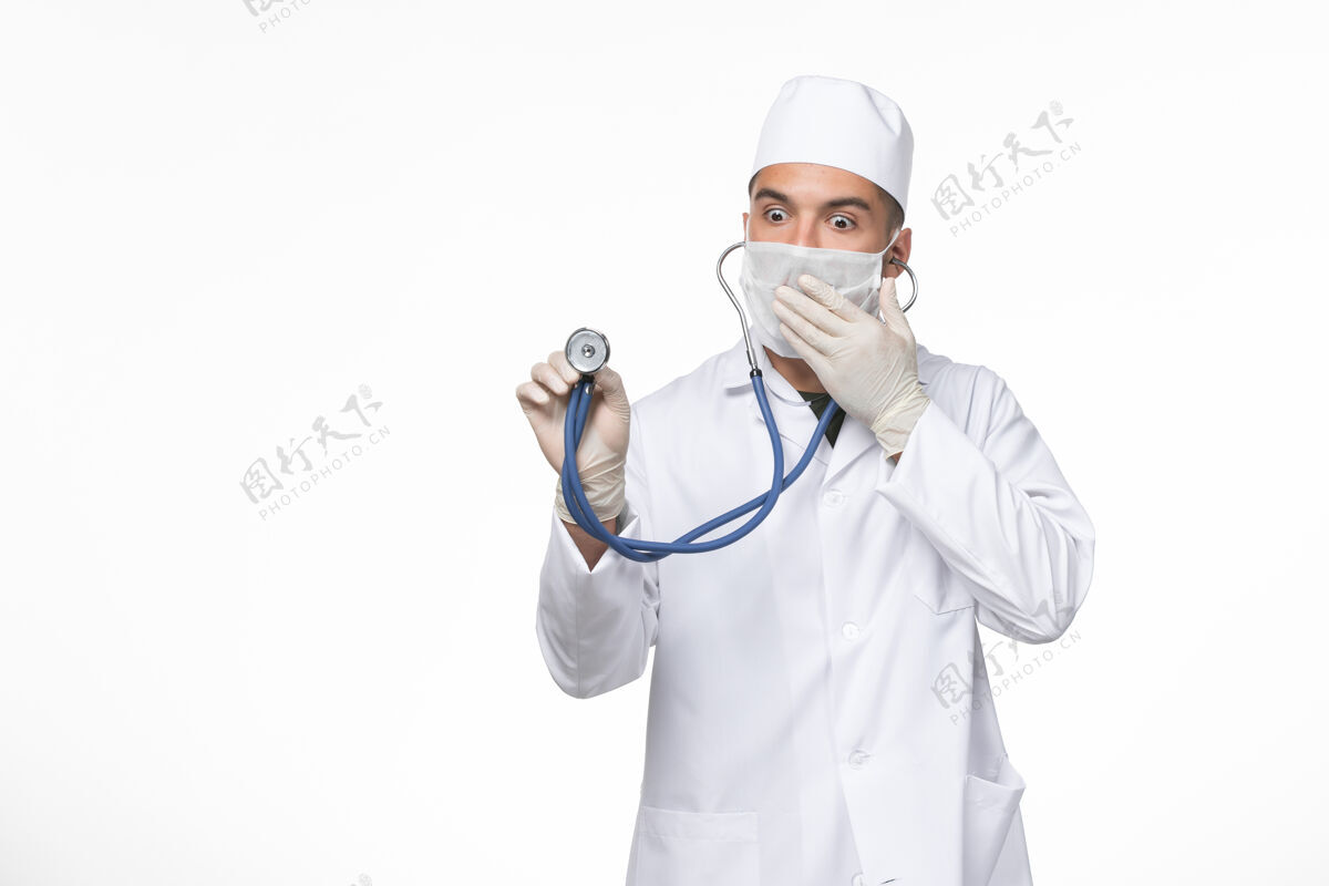 视图正面图：男医生穿着医疗服 戴着口罩 在白色办公桌上用听诊器抵御冠状病毒-冠状病毒疾病大流行病毒男性听诊器