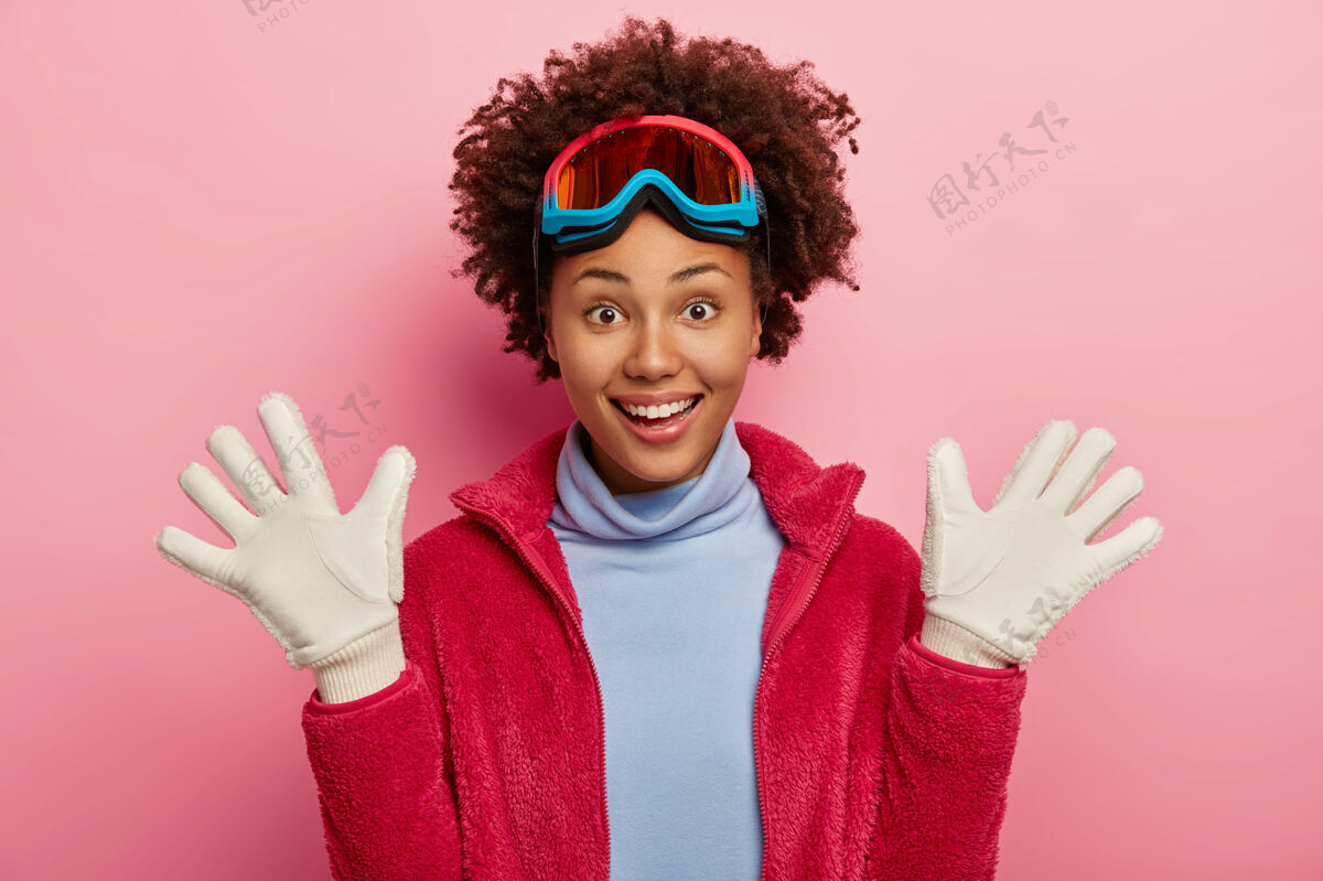 温柔皮肤黝黑的女滑雪者戴着白手套举着手掌 滑雪后玩得很开心 笑得很开心 戴着防护面具 高领毛衣和红色夹克 过着寒假 隔离在粉色背景上请微笑高兴