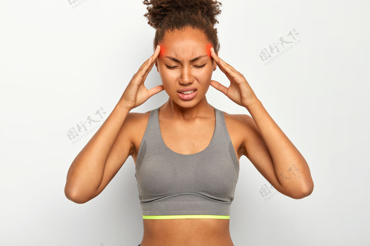 卷曲疲劳过度的美国黑人妇女按摩太阳穴 感到强烈的头痛 闭上眼睛疼痛 患有偏头痛 穿着运动胸罩 紧咬牙齿 隔离在白墙上头压力室内