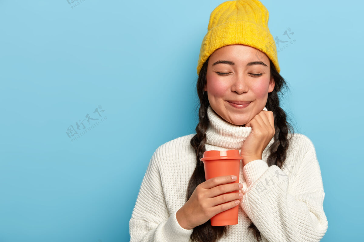 休闲好看的亚洲女人的摄影棚拍摄有两个辫子 戴黄色帽子和白色超大毛衣 拿着外卖咖啡 在蓝色背景下摆姿势 为你的广告复制空间女士外卖自由空间