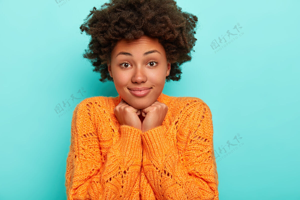惊喜横观兴高采烈好奇惊讶的非洲裔美国女士 深色浓密的头发 双手触碰下巴 穿着鲜艳的橙色毛衣 孤立在蓝色的墙上非洲年轻请