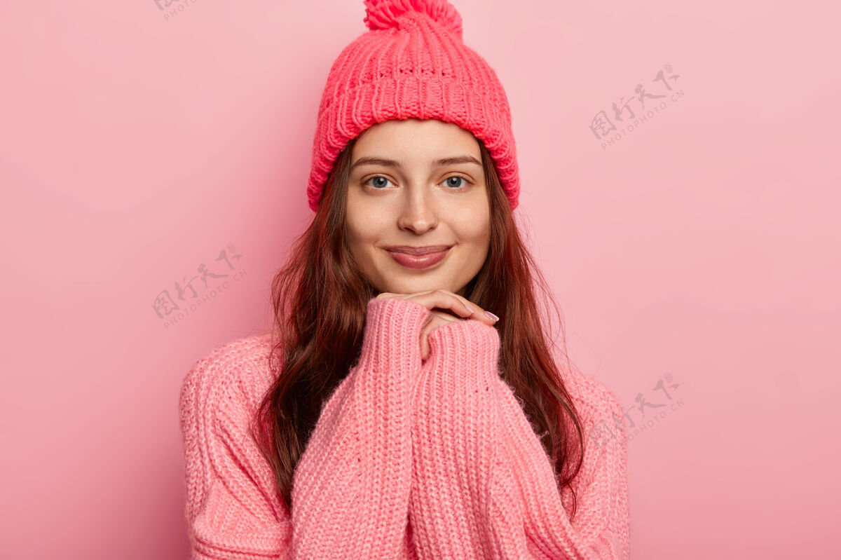 毛衣年轻漂亮的黑发女子双手托着下巴 穿着暖和的冬装 在镜头前愉快地看着有着自然美的不化妆帽子高兴女士