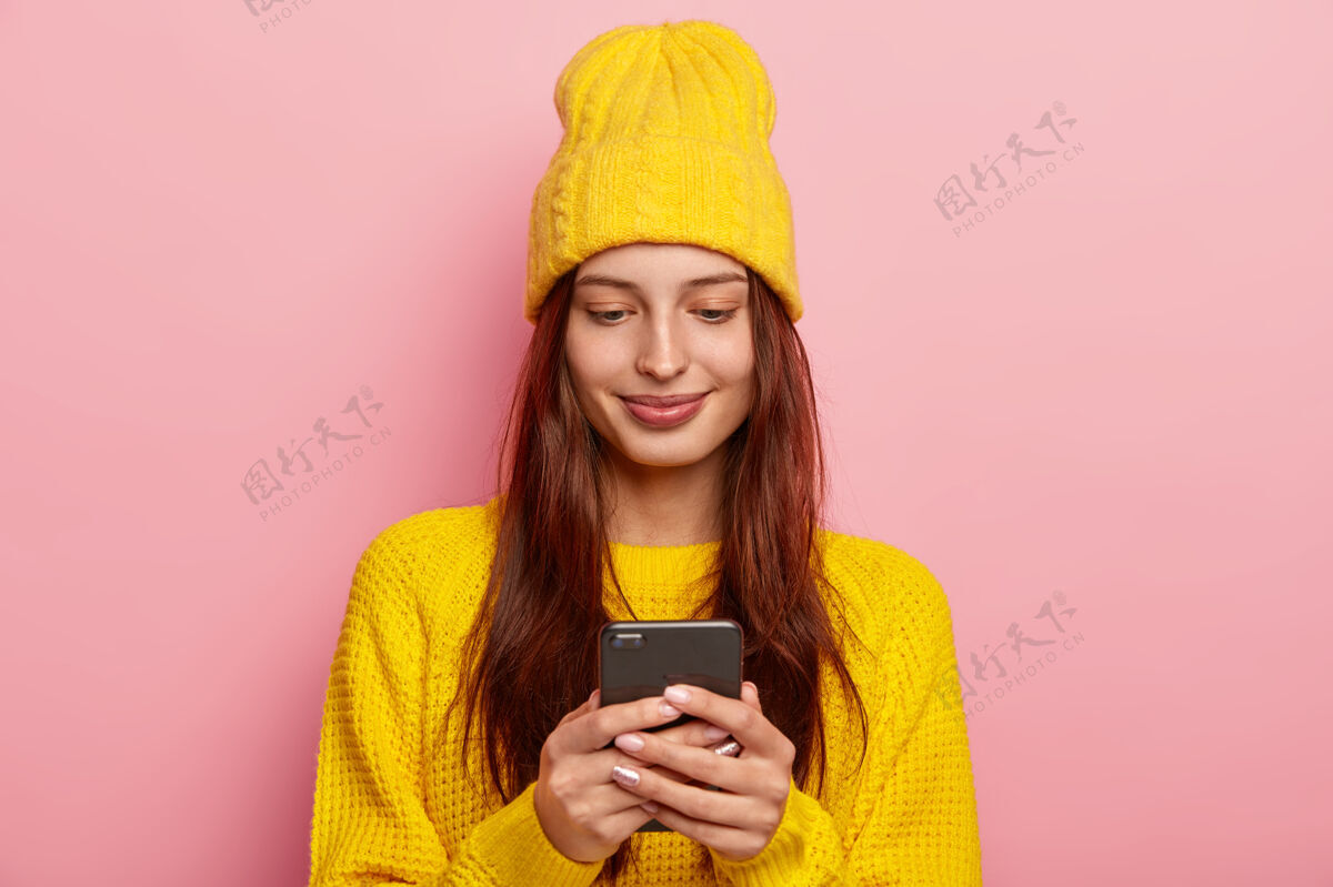 毛衣女性使用智能手机 长直发 穿着黄色毛衣和帽子 有满意的面部表情 在粉色背景下摆姿势表情年轻一个