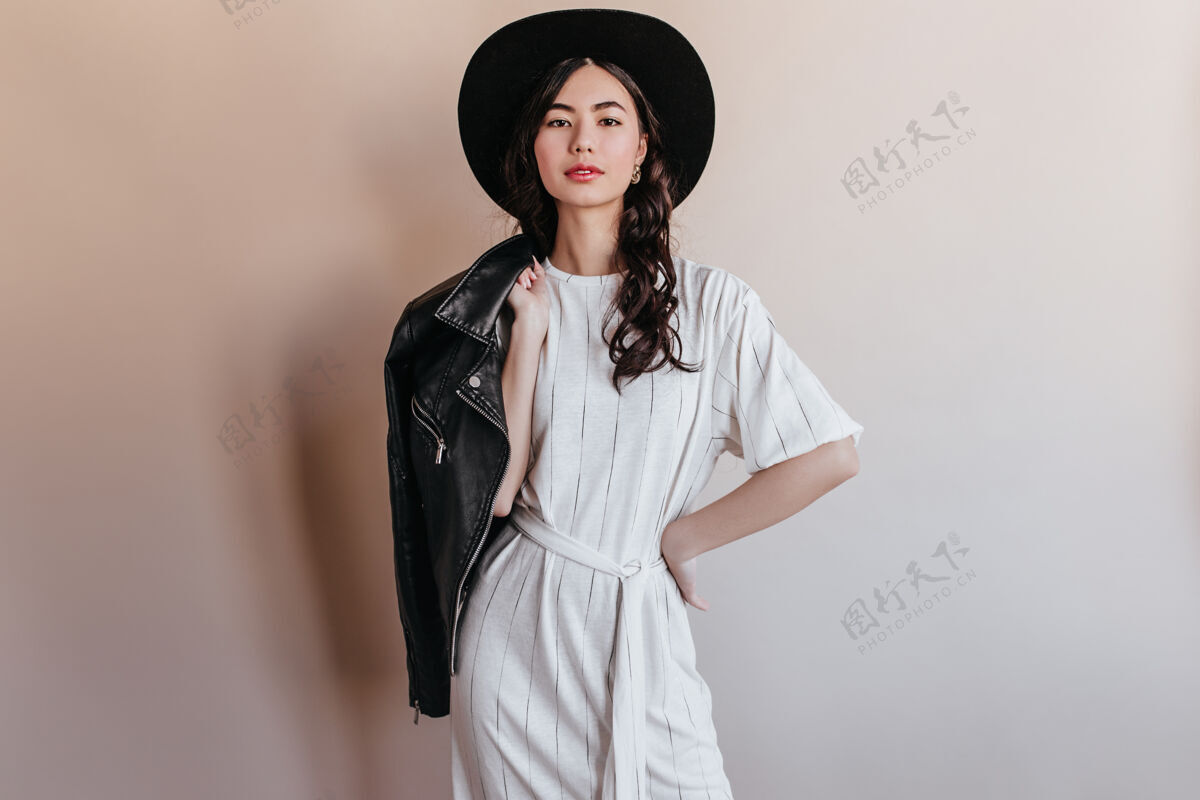 年轻人优雅的亚洲女人穿着白色礼服看着镜头自信的中国女人戴着帽子拿着皮夹克时尚黑发帽子