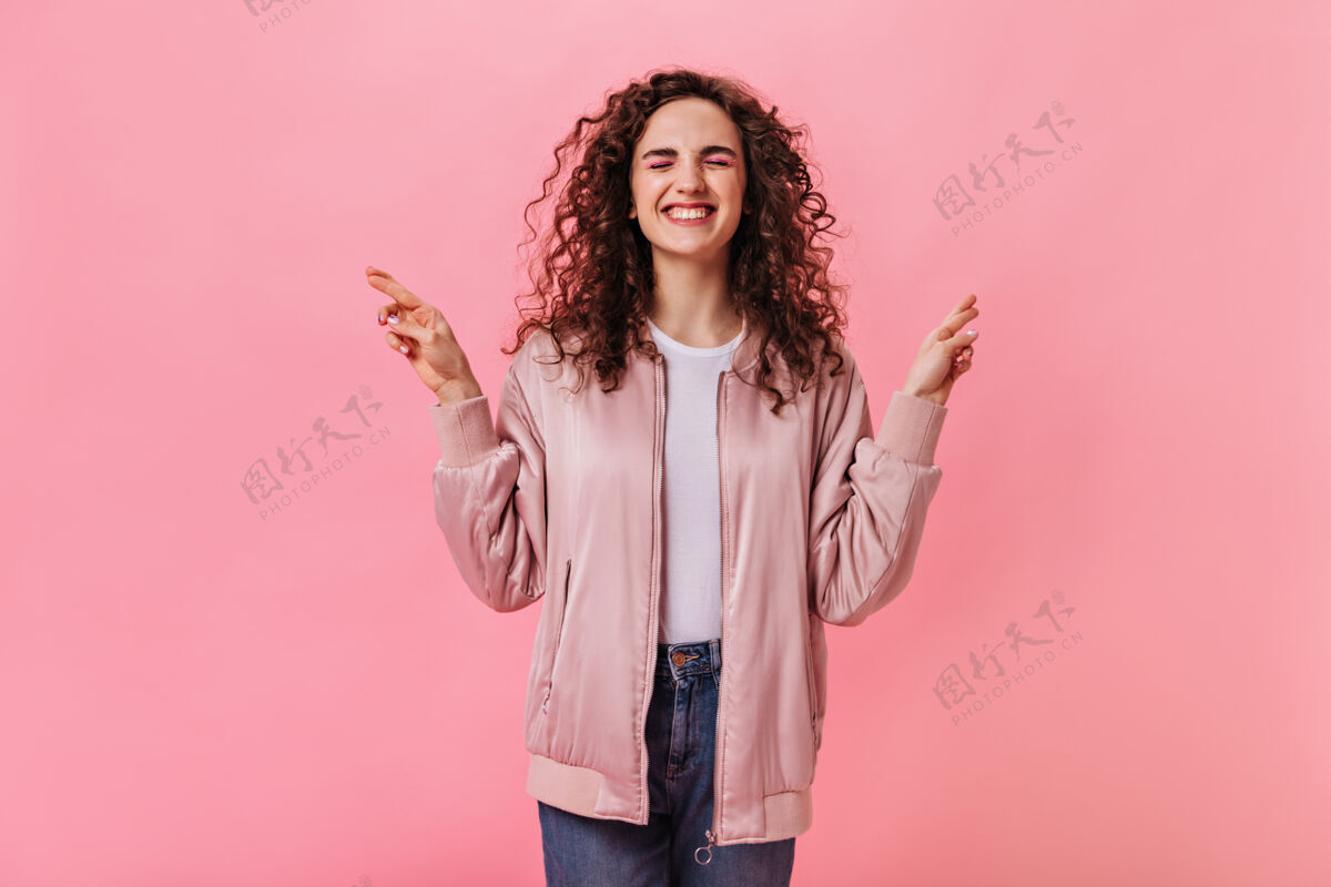 针织穿着时髦夹克的卷发女人交叉着手指衣服粉色背景自信